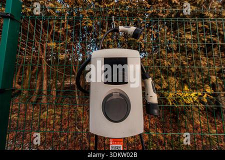 Weathered wall mounted EV charger on a green wire fence with two Type 2 connectors, reflective screen showing hands, and bright orange Obilly QR stick Stock Photo
