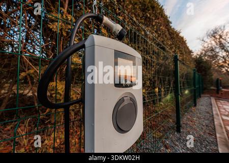 A wall mounted EV charger with coiled cable and Type 2 connector hangs on a green metal fence beside gravel and pavers. Low angle early evening light Stock Photo