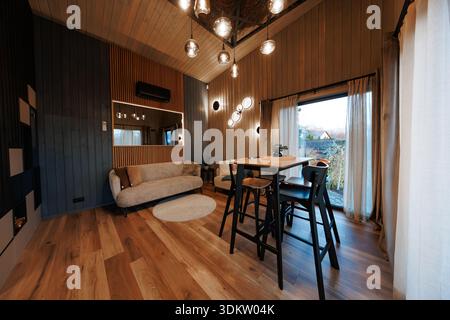 Wood clad room shows compact beige sofa, round rug, high table with four black stools, glass orb pendants, floor to ceiling curtains, sliding doors, a Stock Photo