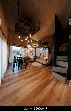 High ceiling loft cabin interior shows tan sofa, glowing wall mirror, cluster pendant lights, tall dining table with stools, sliding doors to garden p Stock Photo