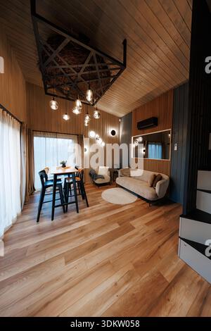 Modern lofted living and dining with geometric twig chandelier focus. Warm wood panels, wide plank floors, pendant bulbs, sofa, bar stools, evening am Stock Photo
