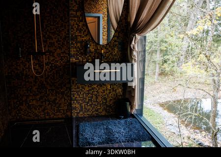 Modern bathroom featuring a dark mosaic tiled wall, floating black vanity with gold accents, oval mirror, and floor to ceiling windows overlooking tre Stock Photo