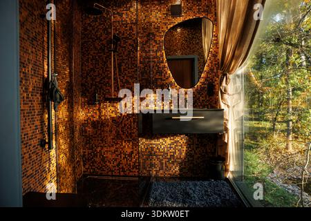 Modern bathroom featuring orange and brown mosaic tiles, a black sink, rainfall shower, and a large window revealing trees and foliage outside. Stock Photo