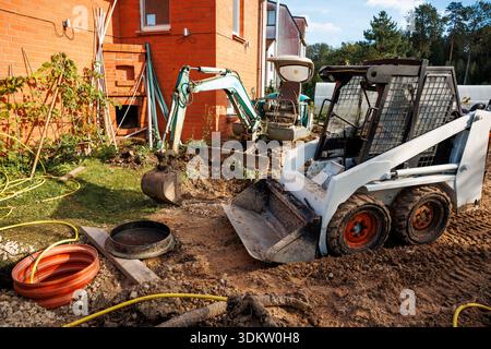 White skid steer loader and green excavator on dirt area near red brick building with vines, surrounded by tools, pipes, and distant trees. Stock Photo