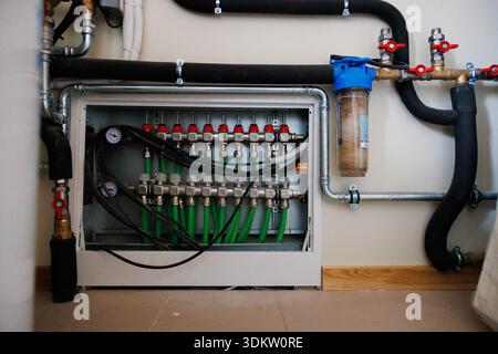 Wall mounted manifold with gauges, red valves, and green PEX lines, plus blue canister filter and insulated pipes, indicating hydronic heating and wat Stock Photo