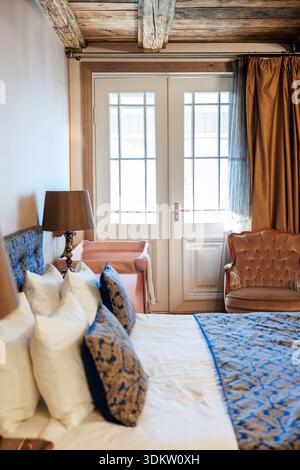 A double bed with patterned blue textiles sits by a brass lamp, velvet armchair, and pink bench before French doors as daylight filters through gold d Stock Photo