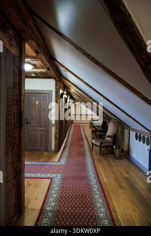 Attic corridor in an old European inn shows exposed beams, red runner, room doors, vintage chairs, and a desk under angled ceilings, lit by warm wall Stock Photo