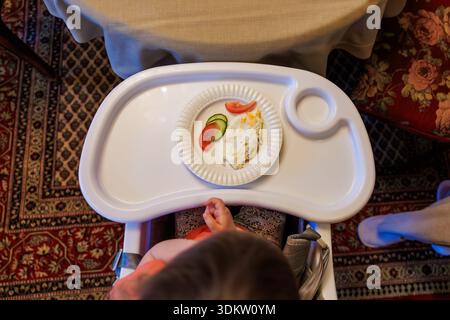 A toddler sits in a white high chair, hands near a paper plate with an omelet, cucumber, and tomato in a carpeted dining room with rugs and soft warm Stock Photo