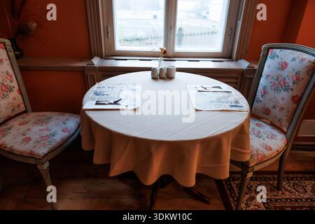 A small round table for two sits by a double hung window with soft daylight, vintage chairs, paper menus, cutlery, condiment shakers, and a single ste Stock Photo