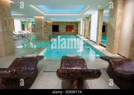 Luxurious indoor spa pool with marble columns and waterfall spout. Submerged steps and lane layout sit under cool recessed lighting with mosaic lounge Stock Photo