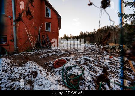 A red brick two story house borders a snow dusted yard with hoses, ladders, pipes, and a white cylindrical tank by trees, in late afternoon with long Stock Photo