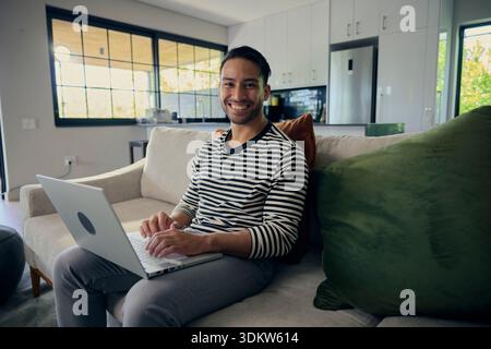 Man comfortably using laptop on couch in house with natural lighting from window Stock Photo