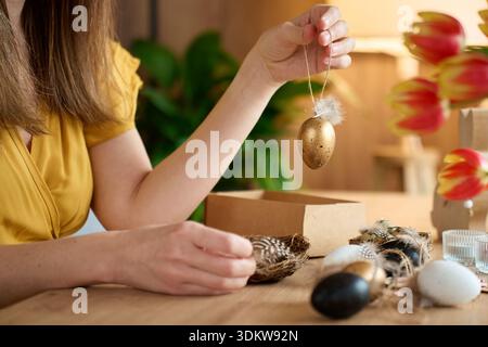 Woman holding decorative golden Easter egg ornament with feather and string over wooden table with black and white eggs and nest Stock Photo