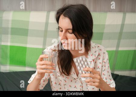 Young woman in pajamas sitting on bed, holding a blister pack of pills and a glass of water, looking down thoughtfully. Concept of illness, recovery, Stock Photo