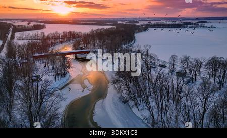 A flock of sandhill cranes fly through this aerial photograph of a river winding through a snowy landscape into the setting sun. Covered bridge center Stock Photo