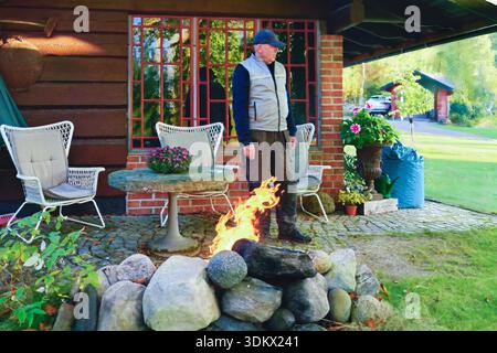 Older man stands by a stone fire pit in a garden, surrounded by comfortable chairs and vibrant plants, creating a serene outdoor ambiance Stock Photo
