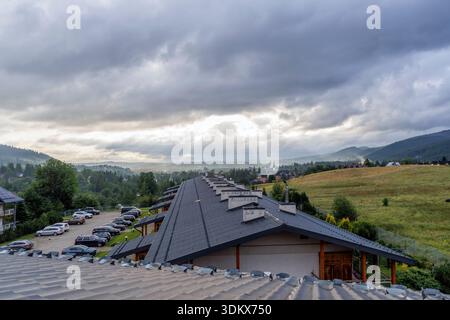 A wide view of a mountain valley resort under a heavy, dramatic sky with sun rays shining on distant hills and a green meadow. Stock Photo