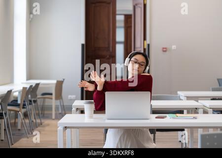 Korean girl stretching hands while watching online webinar for education distant courses or university on laptop in modern coworking space, wear wireless headphones. || Model released Stock Photo