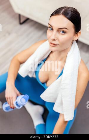 Woman in blue athletic wear sits on the floor with a towel around her neck and a dumbbell, modern living area with light-colored furniture in the back Stock Photo