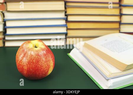 Red apple on the green table and opened books against a background pile of books Stock Photo
