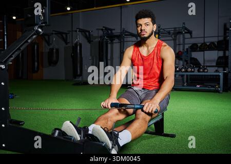 Focused young man with a beard exercises intensely using a rowing machine in the gym. Stock Photo