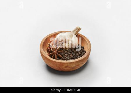 A wooden bowl filled with black peppercorns, star anise, and a whole garlic on a clean white background, highlighting natural textures. Stock Photo