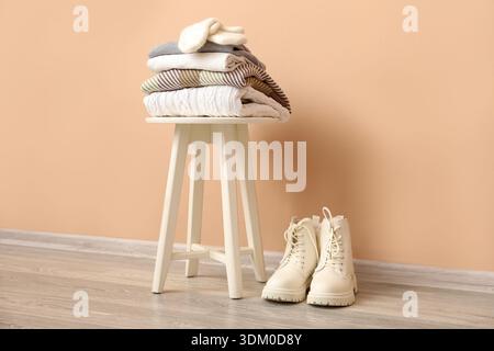 Stack of folded sweaters with gloves on stool and boots near beige wall Stock Photo