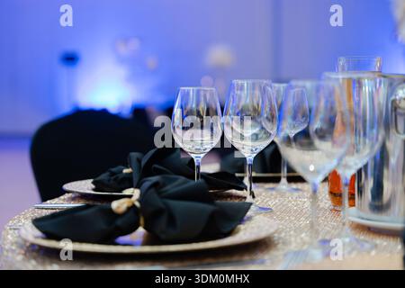 Elegant table setting with crystal wine glasses, black napkins and golden plates in blue lit banquet hall for luxury event or wedding reception Stock Photo