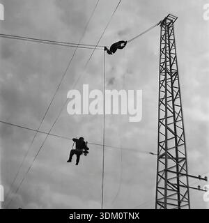 Sloviansk, Donetsk region, Ukraine, USSR. 1960s. A dramatic low-angle archival photograph of two linemen working at height on high-voltage power lines. The men hang suspended on cables near a metal lattice tower. One worker holds insulators while his colleague secures the wire above. This black and white image captures the danger and bravery of the profession during the industrial electrification of the Donbas in the stable and peaceful Soviet era. Stock Photo