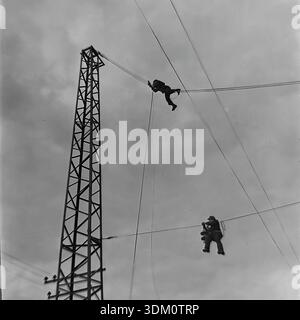 Sloviansk, Donetsk region, Ukraine, USSR. 1960s. A dramatic low-angle archival photograph of two linemen working at height on high-voltage power lines. The men hang suspended on cables near a metal lattice tower. One worker holds insulators while his colleague secures the wire above. This black and white image captures the danger and bravery of the profession during the industrial electrification of the Donbas in the stable and peaceful Soviet era. Stock Photo
