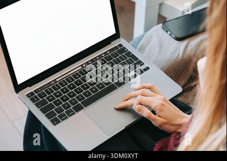 Close-up of a woman using a laptop computer with a blank white screen while sitting at home. A smartphone lies next to the laptop. The image represent Stock Photo