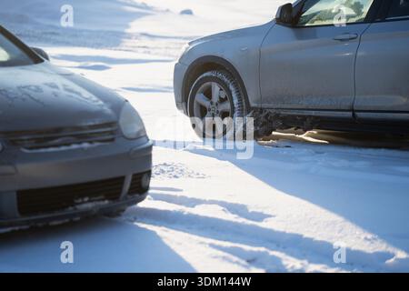 Winter parking lot scene with gray SUV wheel on deep snow, long shadows and sunlight on white ground, blurred car front in foreground showing cold wea Stock Photo