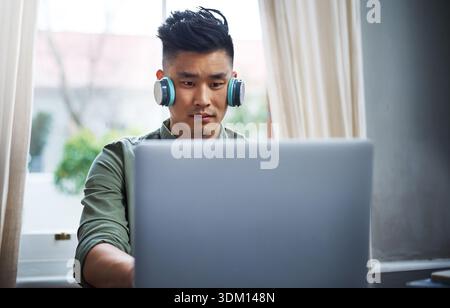 Headphones, reading and asian man with laptop for remote work, system maintenance and feature development. Tech, freelancer or programmer in home for Stock Photo