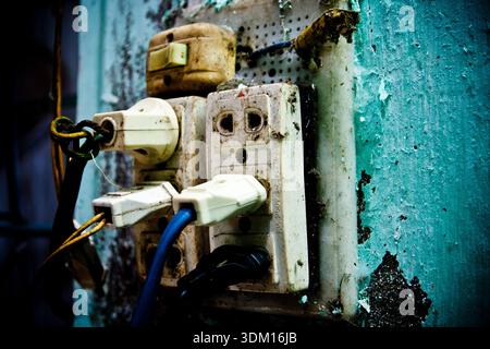 Old electrical outlet with multiple plugs and tangled cables on a worn wall inside a traditional shoemaker’s workshop in Vietnam, highlighting unsafe Stock Photo