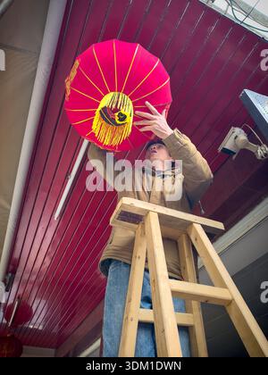 A skilled senior Asian man carefully hangs a bright red lantern on a ceiling, showcasing the dedication and craftsmanship involved in creating a festi Stock Photo