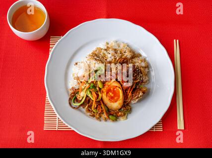 Glazed pork belly with rice, salad coleslaw and peanut (kung pao style) on white plate, chopsticks and tee on red background. Stock Photo