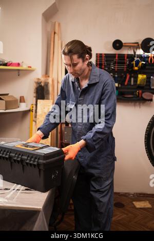 Professional repairman in workwear holding toolbox standing in garage workshop with tools. Concept of industry, transport maintenance Stock Photo