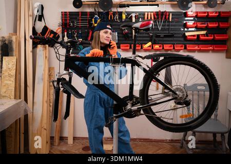 repair woman in bicycle shop, cycling workshop. Female bike mechanic repairing bicycle speaking on smartphone during work. start-up, small business ow Stock Photo