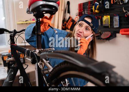 repair woman in bicycle shop, cycling workshop. Female bike mechanic repairing bicycle speaking on smartphone during work. start-up, small business ow Stock Photo