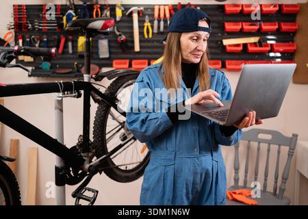 Small business owner using a laptop for bookkeeping in her bike shop. Female entrepreneur managing repair records and inventory on a computer in a gar Stock Photo