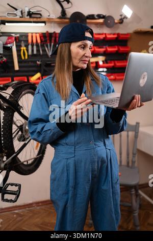 Female bike mechanic multitasking inspecting and repairing bike using laptop computer browsing the internet in bicycle repair workshop. copy space Stock Photo