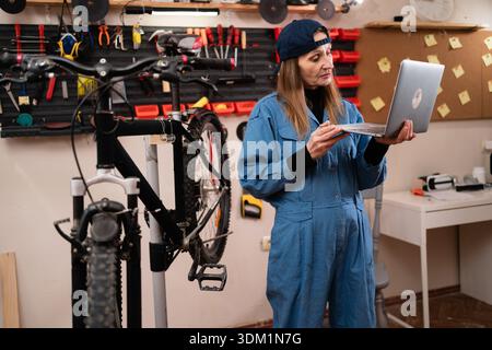 Female bike mechanic multitasking inspecting and repairing bike using laptop computer browsing the internet in bicycle repair workshop. copy space Stock Photo