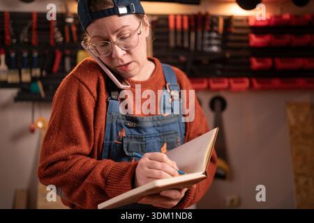 A professional carpenter works with wood using carpentry tools in her garage, taking orders over the phone and writing them down in a notebook. Concep Stock Photo