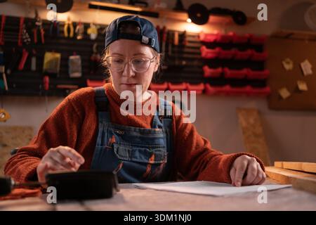 A professional female carpenter works with wood using carpentry tools in her garage, sitting and calculating on a calculator. Concept of profession, a Stock Photo