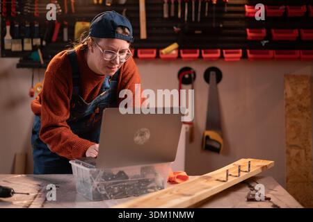 A professional female carpenter works with wood using carpentry tools in her garage. She stands at a table and types on a laptop keyboard. Conceptuali Stock Photo