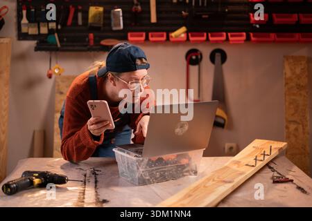 A professional female carpenter works with wood using carpentry tools in her garage. She holds a smartphone and searches online using a laptop. Concep Stock Photo