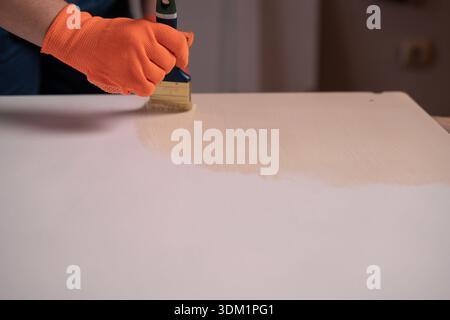 Female hand in orange glove with paintbrush painting on wooden part of table. Stock Photo