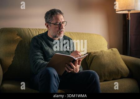 A middle-aged man sits on a green sofa in his living room, thoughtfully writing in a notebook to practice mindfulness and mental clarity during a quie Stock Photo
