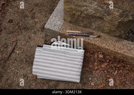 A white and black striped bag is laying on the ground next to a stone pillar. A pair of paintbrushes and a tube of blue paint are also on the ground Stock Photo