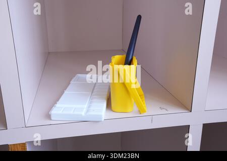A yellow bucket with a black brush sits on a shelf next to a white palette. The scene suggests that someone is preparing to paint or draw Stock Photo